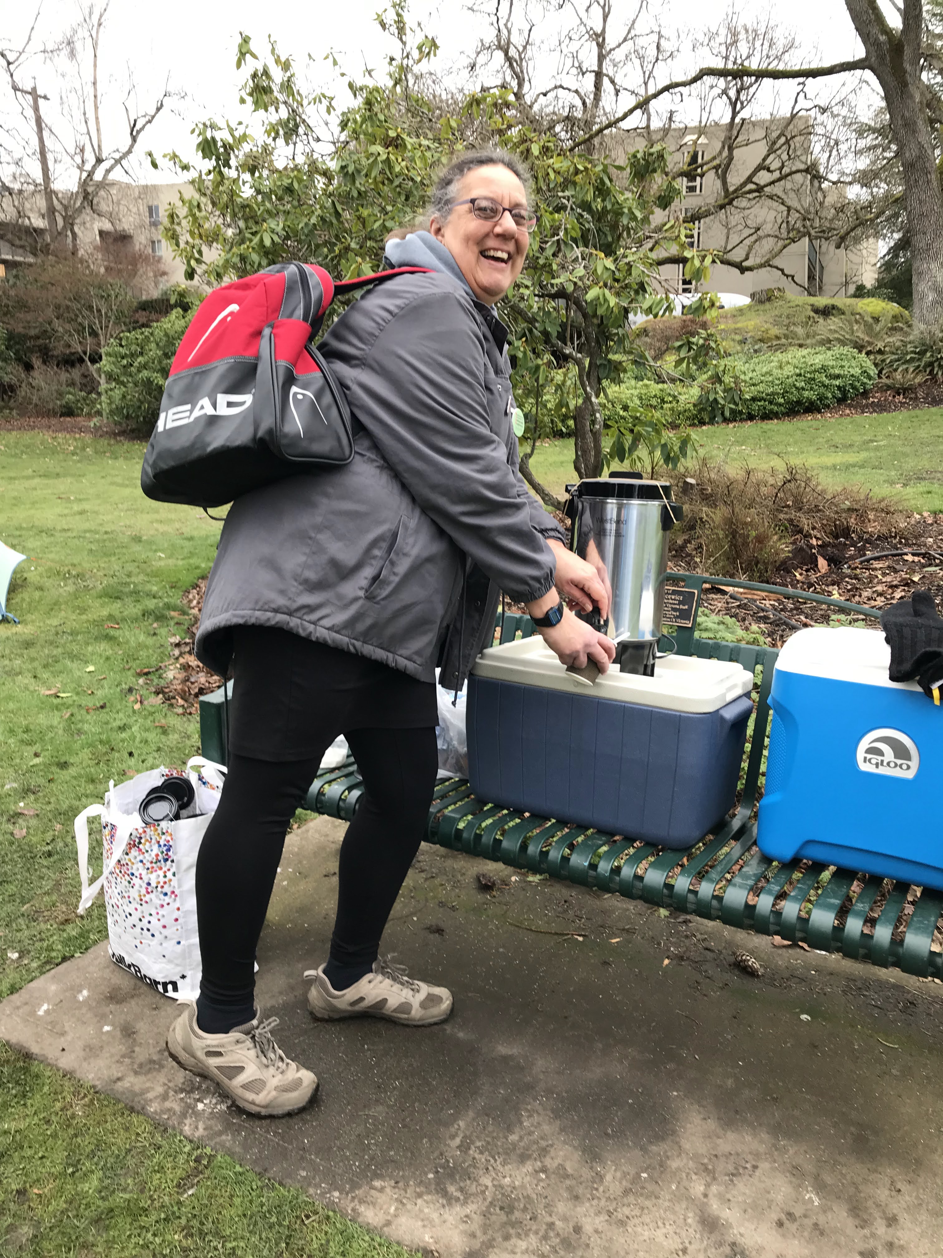 A woman in a rain jacket and backpack dispensing a cup of coffee from an urn in Stadacona Park in Victoria, British Columbia - February 2023